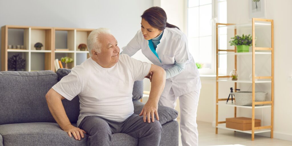 Female caregiver in white scrubs assisting a senior man rising from a couch, illustrative of elder care and Florida guardianship law.